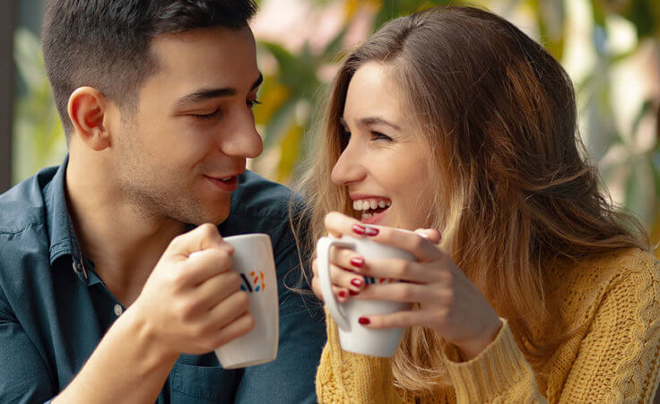 a young man and woman smile at each while holding coffee mugs