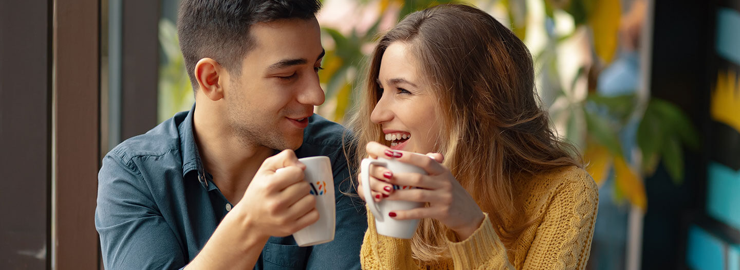 a young man and woman smile at each while holding coffee mugs