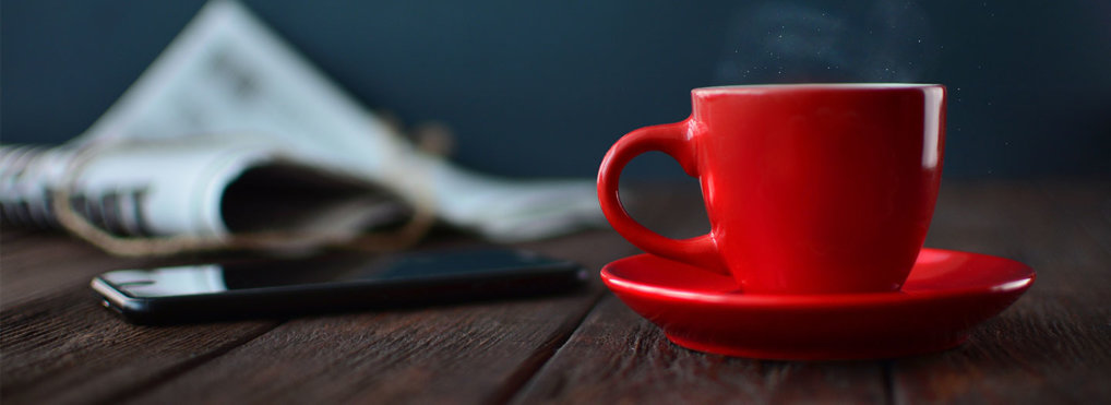 Steam rises from a red coffee mug on a dark wood table. A phone and newspaper lay in the background.