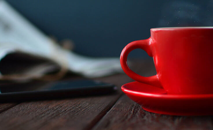 Steam rises from a red coffee mug on a dark wood table. A phone and newspaper lay in the background.