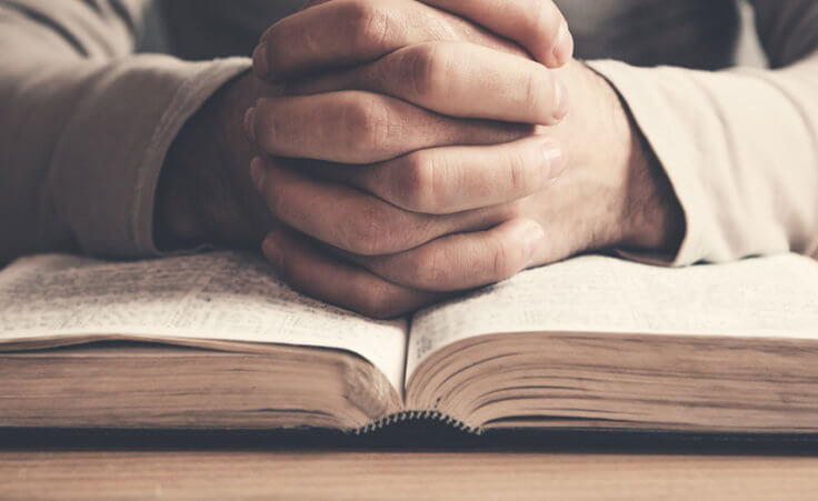A man folds his hands in prayer on top of an open Bible