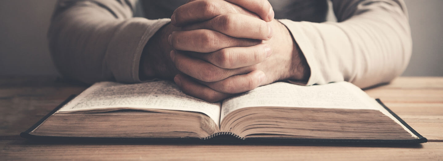 A man folds his hands in prayer on top of an open Bible
