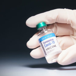 A gloved hand holds a vial labeled COVID-19 vaccine