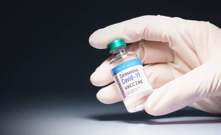 A gloved hand holds a vial labeled COVID-19 vaccine