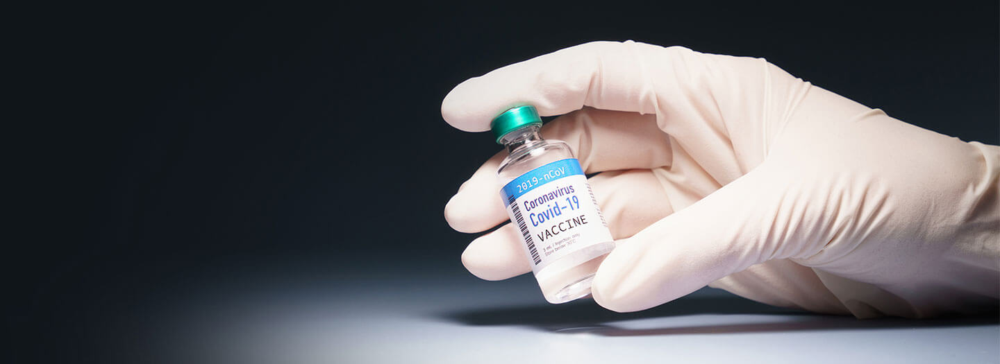 A gloved hand holds a vial labeled COVID-19 vaccine