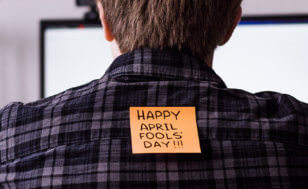 An orange post-it reading "Happy April Fools' Day" is stuck to the back of a man looking at a computer screen