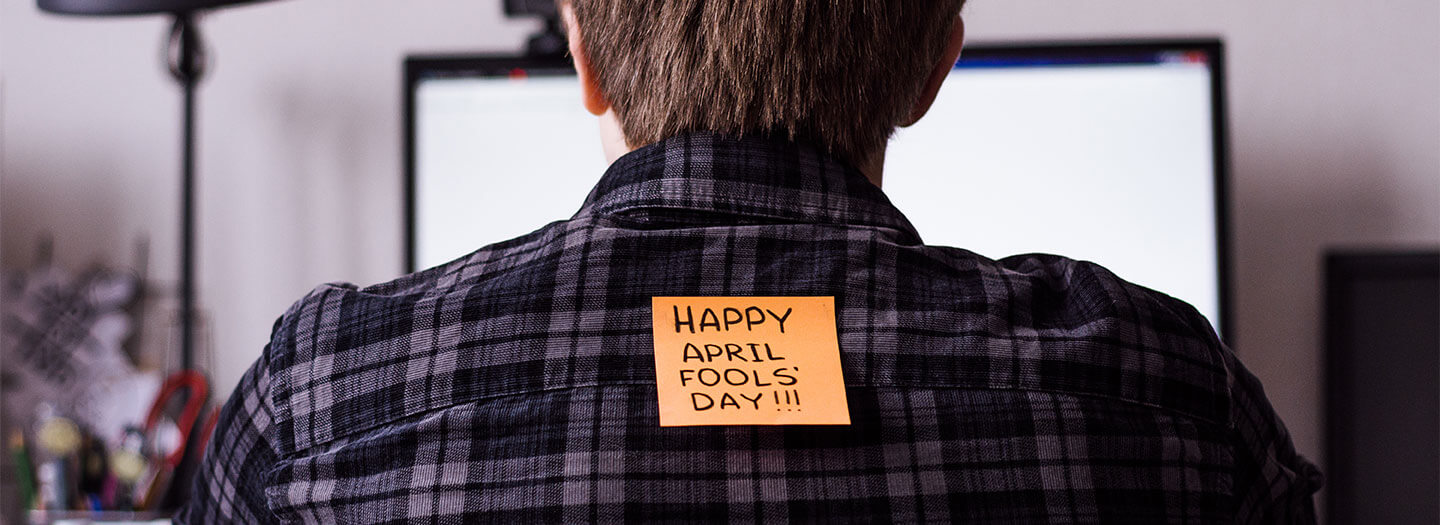 An orange post-it reading "Happy April Fools' Day" is stuck to the back of a man looking at a computer screen
