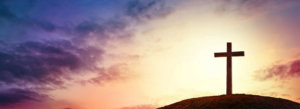 A lone cross sits atop a hill in front of a multihued sky