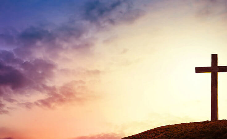 A lone cross sits atop a hill in front of a multihued sky