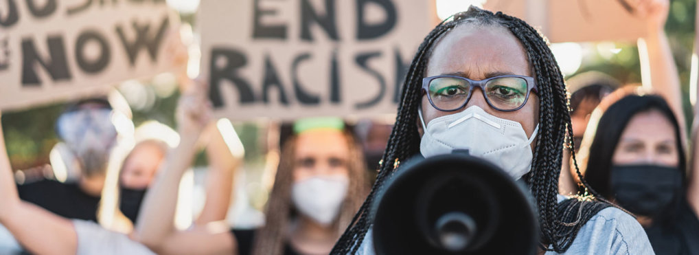 A woman wearing a face mask holds a megaphone toward the camera, leading a social justice protest against racism