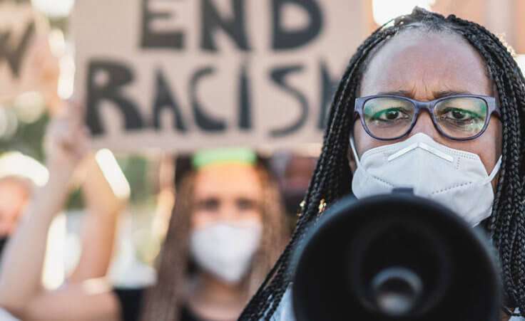 A woman wearing a face mask holds a megaphone toward the camera, leading a social justice protest against racism