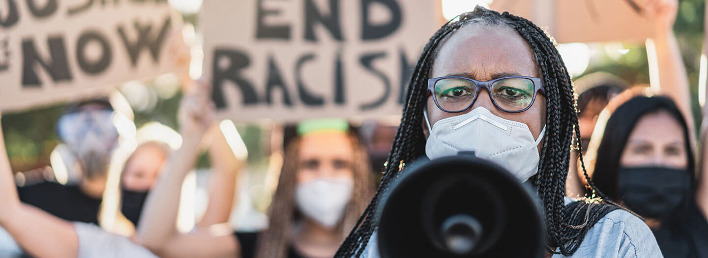 A woman wearing a face mask holds a megaphone toward the camera, leading a social justice protest against racism