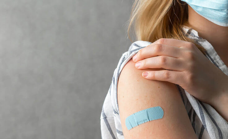 A woman wearing a blue face mask holds her sleeve up on her right arm to reveal a blue bandage, representing having taken the COVID-19 vaccine