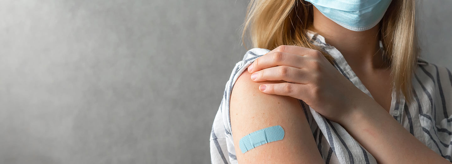 A woman wearing a blue face mask holds her sleeve up on her right arm to reveal a blue bandage, representing having taken the COVID-19 vaccine