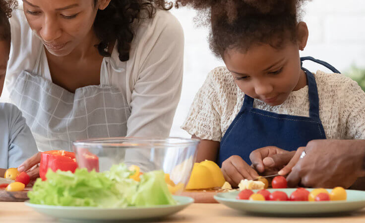 A mother and father with their daughters cut vegetables on a table