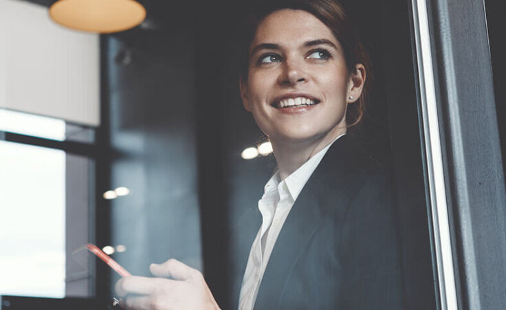 A businesswoman looks out of an office window while holding her cellphone