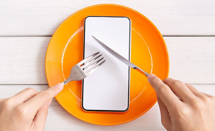 Social media fast: A person pretends to cut into a smartphone on an orange plate using a fork a knife