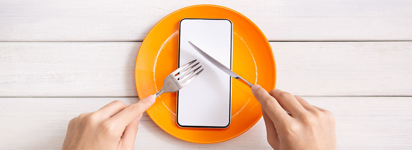 Social media fast: A person pretends to cut into a smartphone on an orange plate using a fork a knife