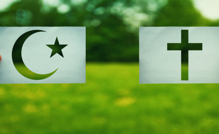 Two hands holding different faith symbols, Islam vs Christianity belief over green field nature.