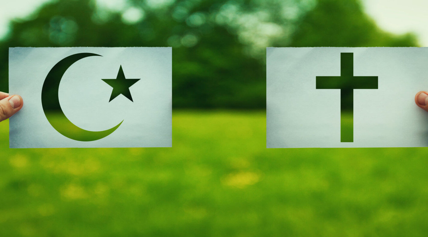Two hands holding different faith symbols, Islam vs Christianity belief over green field nature.