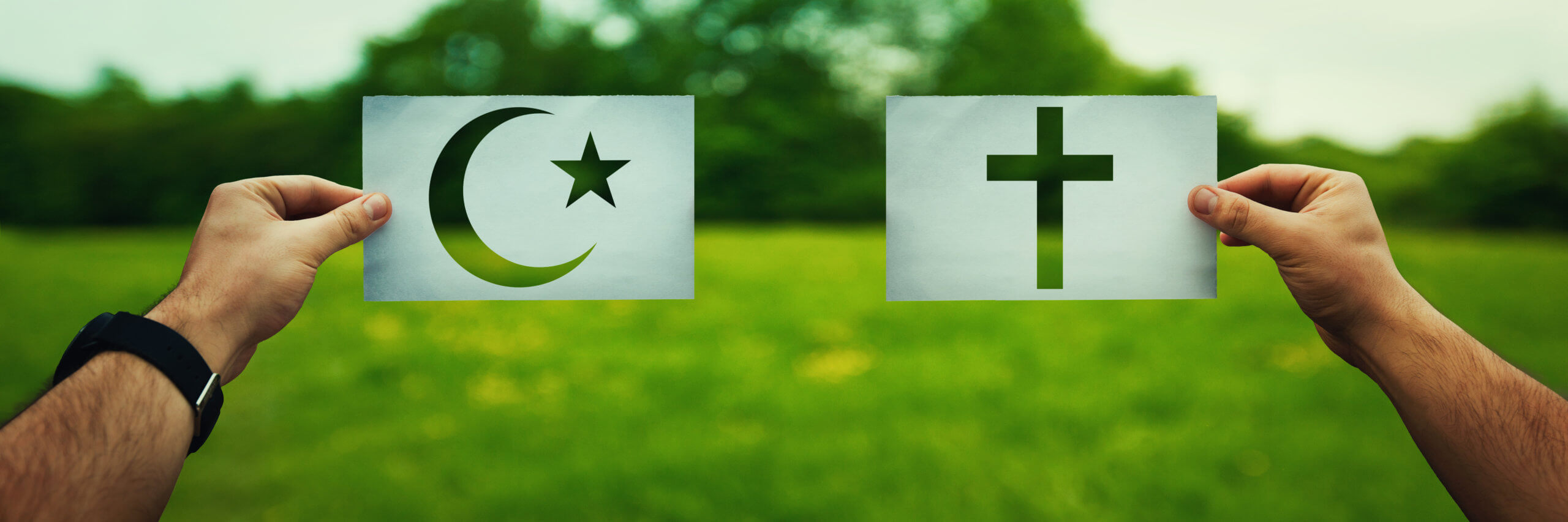 Two hands holding different faith symbols, Islam vs Christianity belief over green field nature.