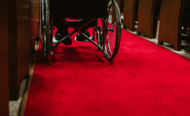 The feet of a disabled man rest on the footsteps of his wheelchair, which sits on a red carpet in the middle between church pews © MIA Studio