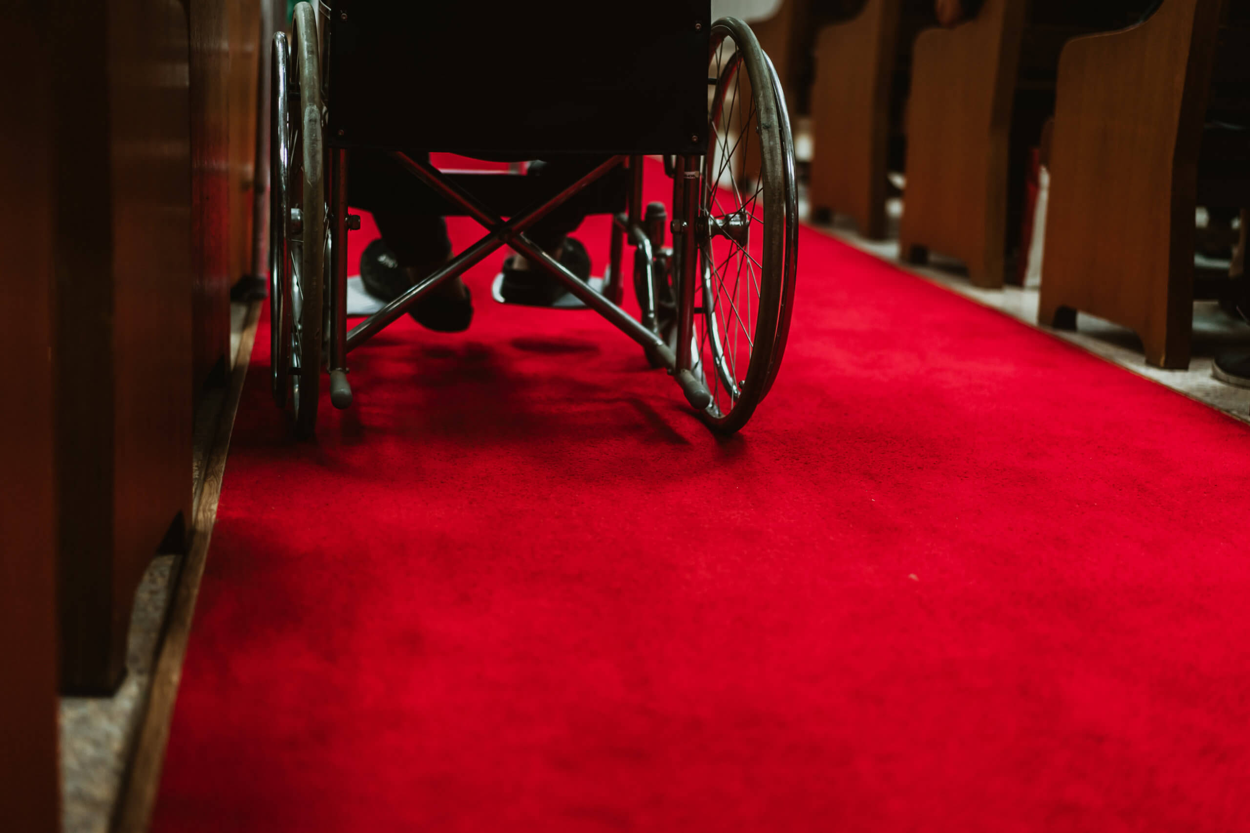 The feet of a disabled man rest on the footsteps of his wheelchair, which sits on a red carpet in the middle between church pews © MIA Studio