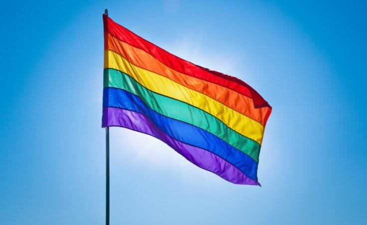 A gay pride flag flies in front of a blue sky. © Alexander Demyanenko/stock.adobe.com
