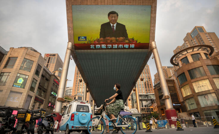 A woman wearing a face mask rides a bicycle past a large television screen at a shopping center displaying Chinese state television news coverage of Chinese President Xi Jinping's visit to Hong Kong in Beijing, Friday, July 1, 2022. (AP Photo/Mark Schiefelbein)