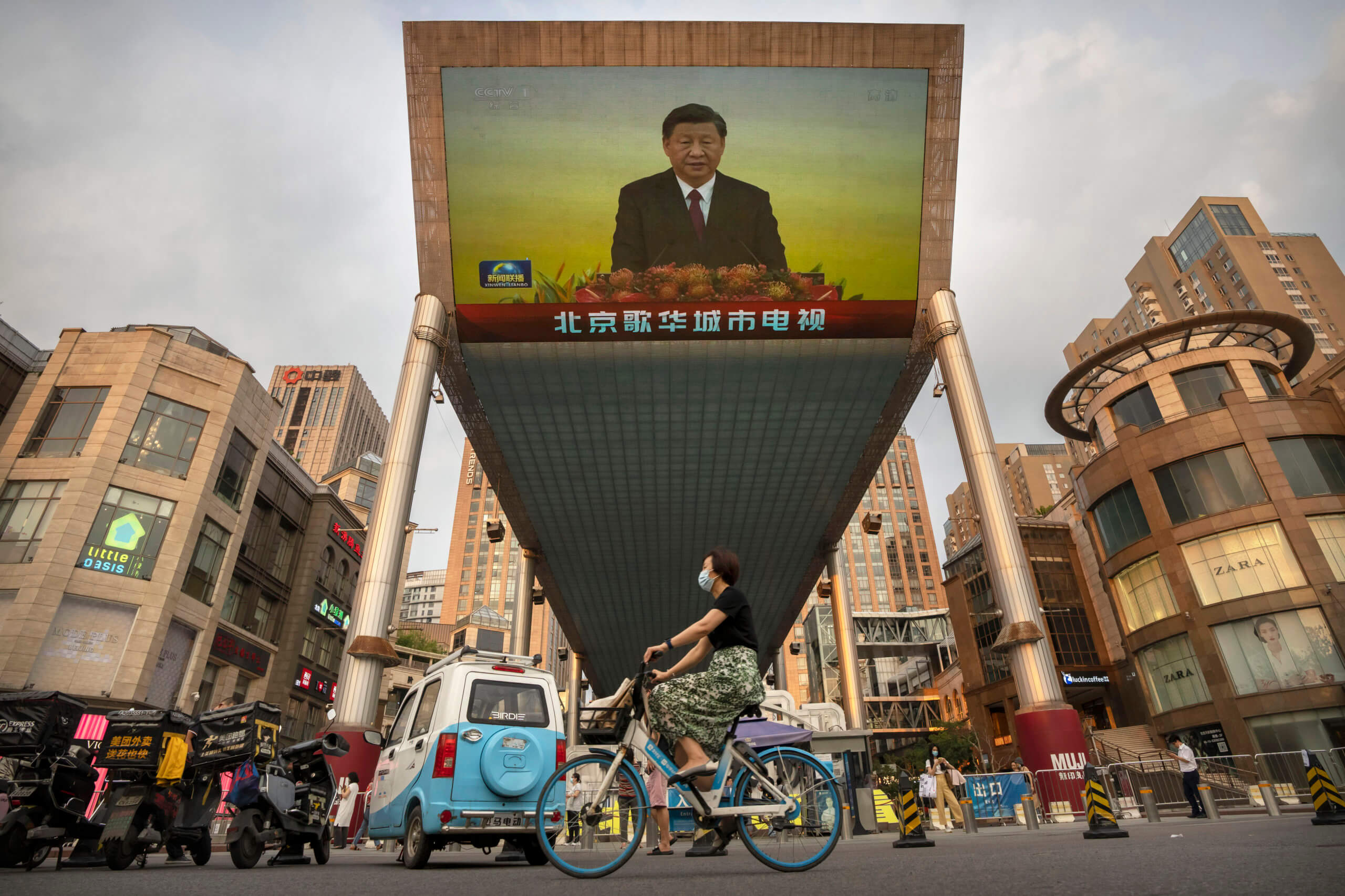 A woman wearing a face mask rides a bicycle past a large television screen at a shopping center displaying Chinese state television news coverage of Chinese President Xi Jinping's visit to Hong Kong in Beijing, Friday, July 1, 2022. (AP Photo/Mark Schiefelbein)