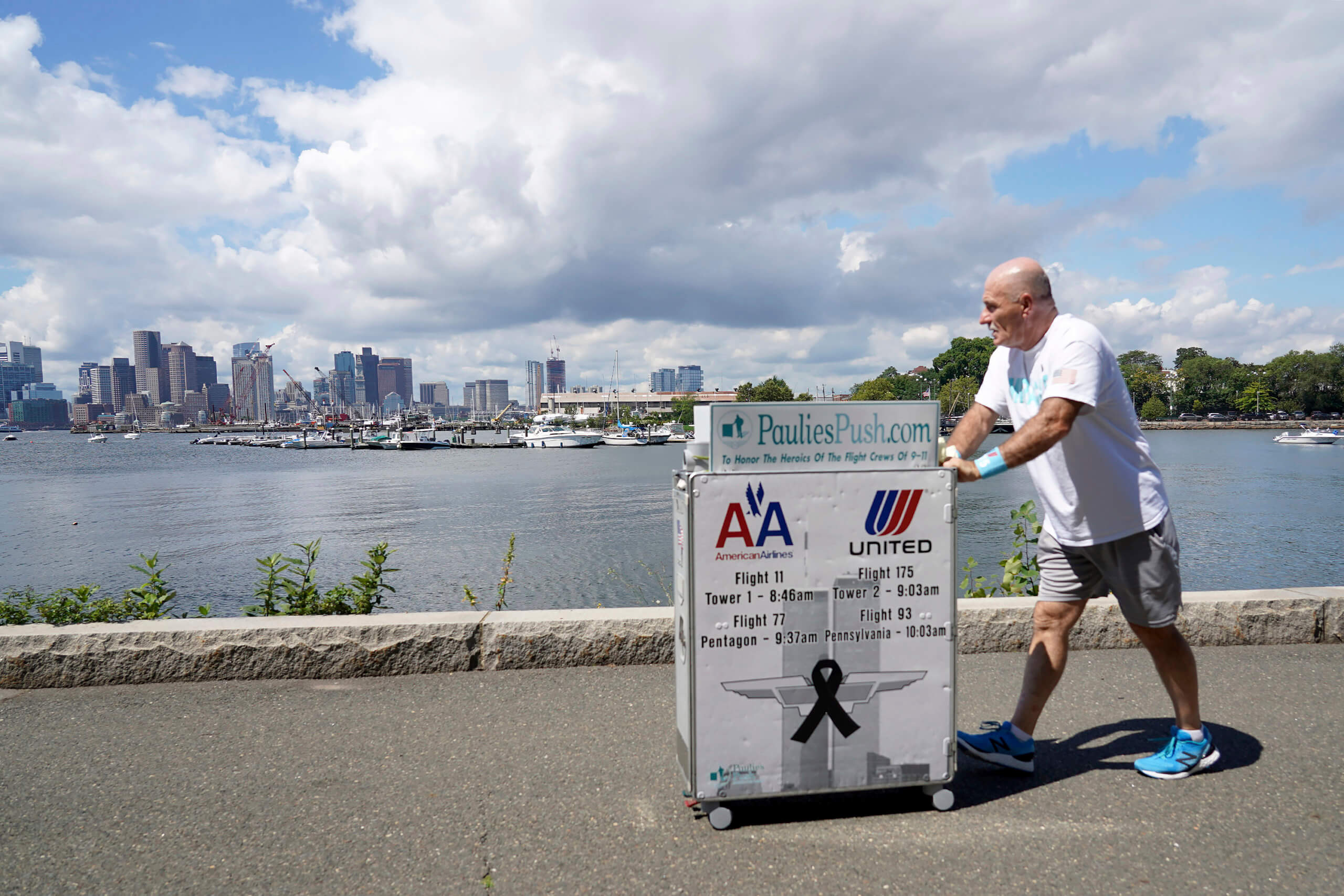 Paul Veneto pushes a beverage cart along the Boston harbor, Saturday, Aug. 21, 2021. Veneto, a former flight attendant who lost several colleagues when United Flight 175 was flown into the World Trade Center on Sept. 11, 2001, is honoring his friends on the 20th anniversary of the terrorist attacks by pushing the beverage cart from Boston to ground zero in New York. (AP Photo/Mary Schwalm)