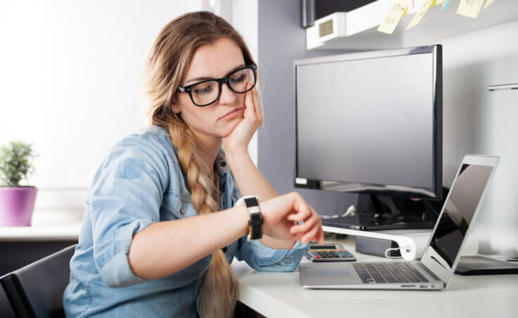 Stock photo: A woman at work checks her watch while sitting in front of her computer. Is she quiet quitting? © leszekglasner /stock.adobe.com