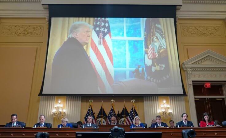The Jan. 6 committee meets with a projected image of then-president Donald Trump behind and above them.