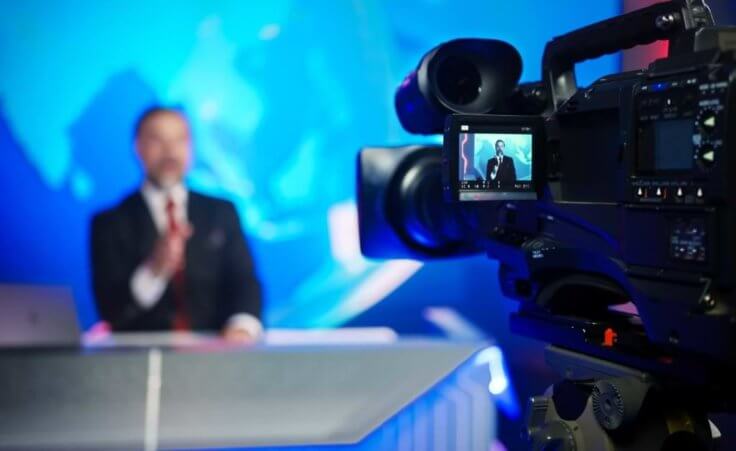 A news anchor talks into a professional TV camera in a studio setting. © Gorodenkoff/stock.adobe.com