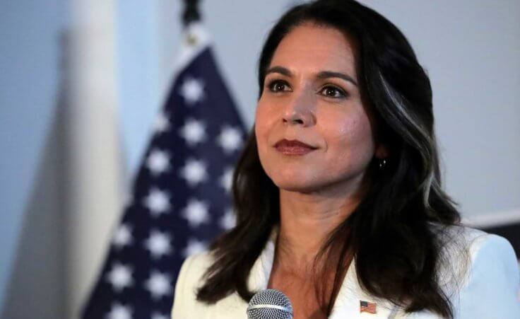 A closeup of Democratic presidential candidate Tulsi Gabbard wearing a white suit in front of an American flag in 2019