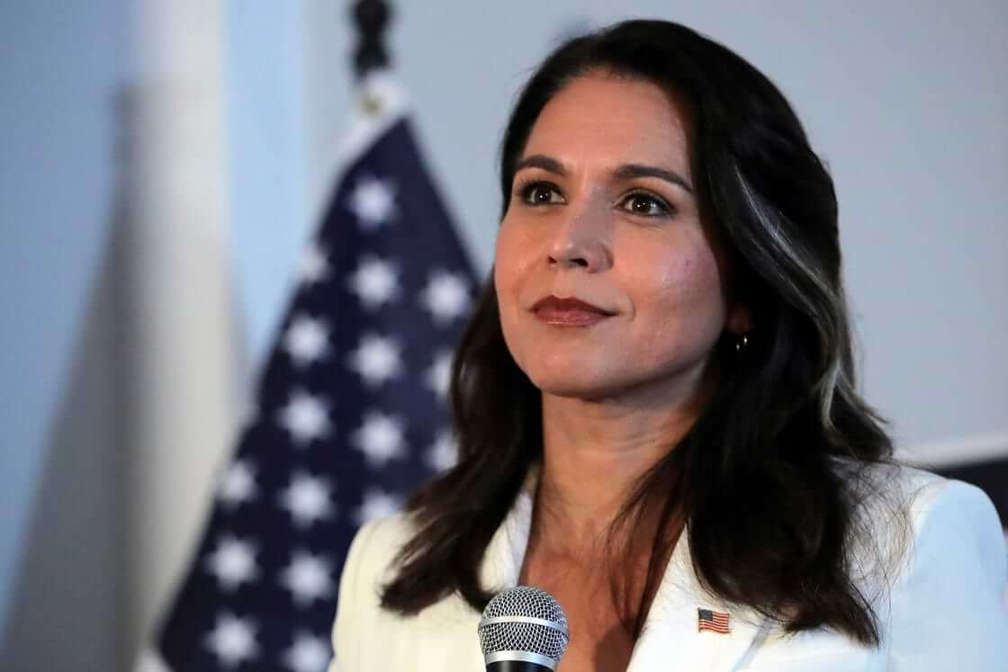 A closeup of Democratic presidential candidate Tulsi Gabbard wearing a white suit in front of an American flag in 2019