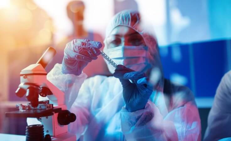 Next to a microscope in the foreground, a vaccine researcher in a clean suit inspects a vial. © alphaspirit/stock.adobe.com