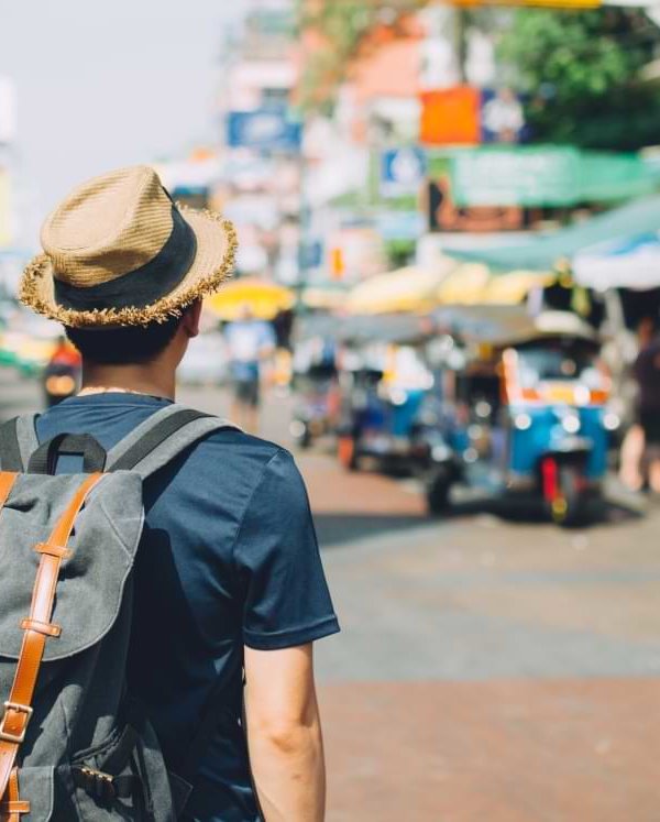 A young man in a straw hat wears a backpack while looking at a busy marketplace in Thailand. © twinsterphoto