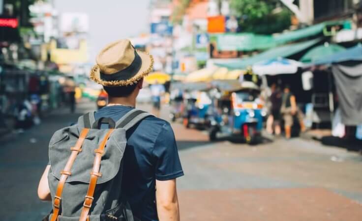 A young man in a straw hat wears a backpack while looking at a busy marketplace in Thailand. © twinsterphoto