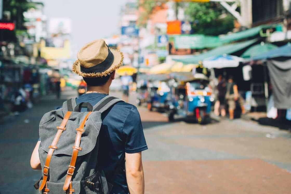 A young man in a straw hat wears a backpack while looking at a busy marketplace in Thailand. © twinsterphoto
