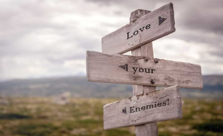 Three wooden signs with arrows read "Love your enemies" on a deserted road.