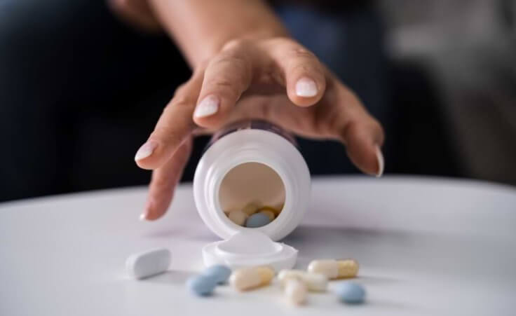 A woman's hand stretches toward an open pill bottle with depression medication spilled onto the table.