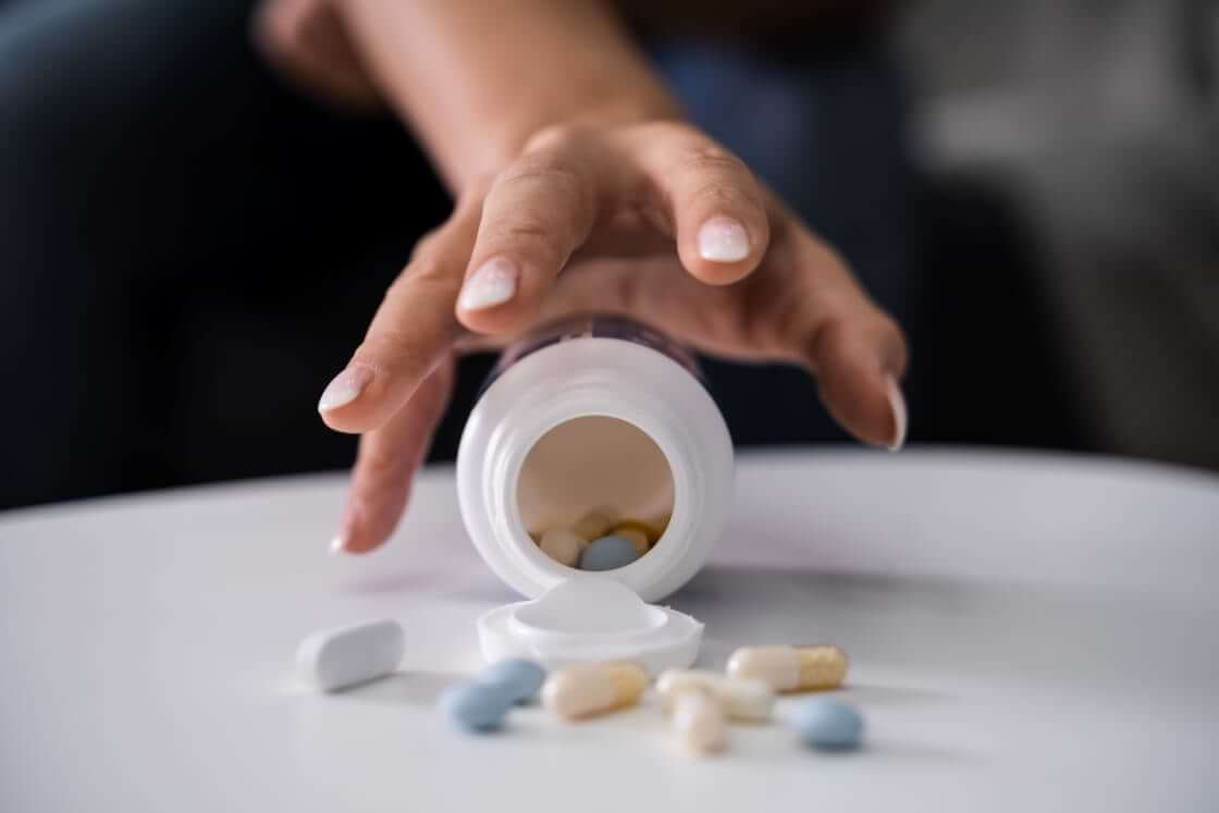 A woman's hand stretches toward an open pill bottle with depression medication spilled onto the table.