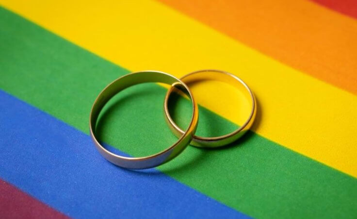 Two overlapping wedding rings sit on top of an LGBTQ pride rainbow background