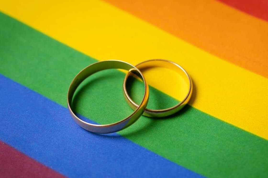Two overlapping wedding rings sit on top of an LGBTQ pride rainbow background