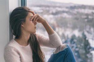 A woman sits next to a window overlooking a winter landscape, her hand covering her eyes in a depressed state.