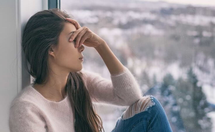 A woman sits next to a window overlooking a winter landscape, her hand covering her eyes in a depressed state.