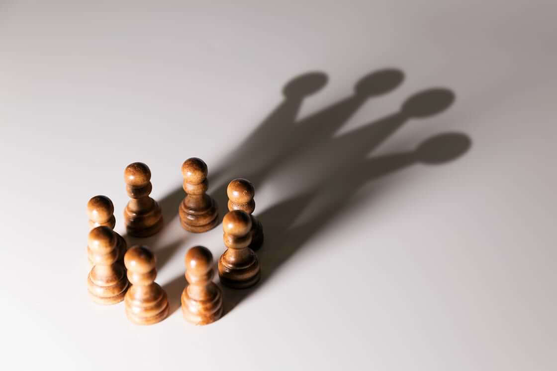 Eight wooden pawn pieces form a circle, casting a singular shadow of a crown onto a white background