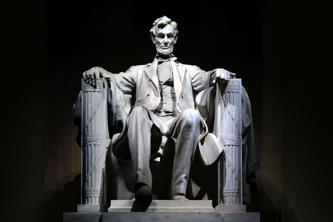 Light shines upon the Abraham Lincoln memorial statue against a black background.