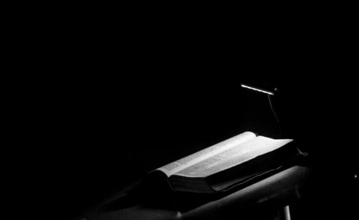 In a black-and-white image, an open Bible sits on a lectern, a single light shining down on it.